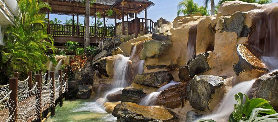 Ein Wasserfall im Tabaiba Princess Hotel mit einer hölzernen Brücke und Pflanzen im Hintergrund.