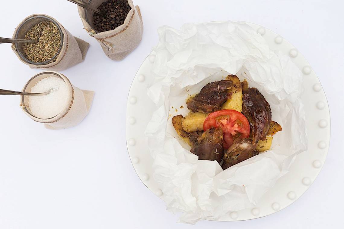 Ein Teller mit gewürztem Fleisch, Tomaten und Kartoffeln, alles in weißem Papier eingewickelt, neben zwei Stoffbeuteln mit Gewürzen.