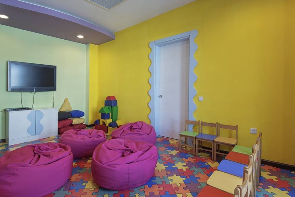 Kinderspielzimmer im Alba Queen Hotel mit bunten Kissen, Spielzeug und einem Fernseher an der Wand.