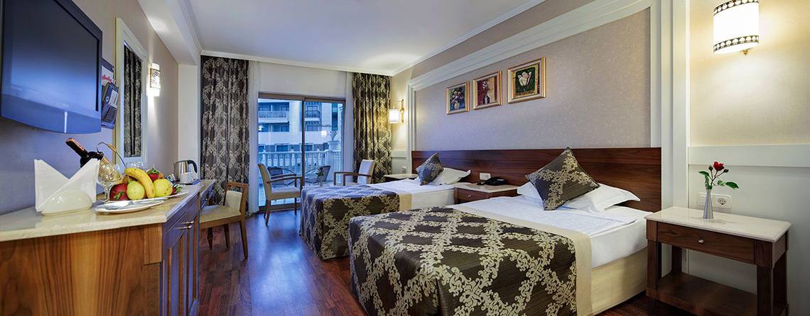 Innenansicht eines Hotelzimmers im Alba Queen mit zwei Betten, einem Fernseher, einem Tisch mit Obst und einem Nachttisch mit einer Vase.