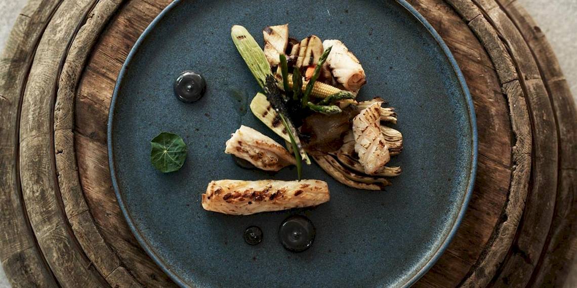 Ein Gericht auf einem blauen Teller mit Holzuntergrund, bestehend aus gegrilltem Fisch, Gemüse und schwarzen Soßen.