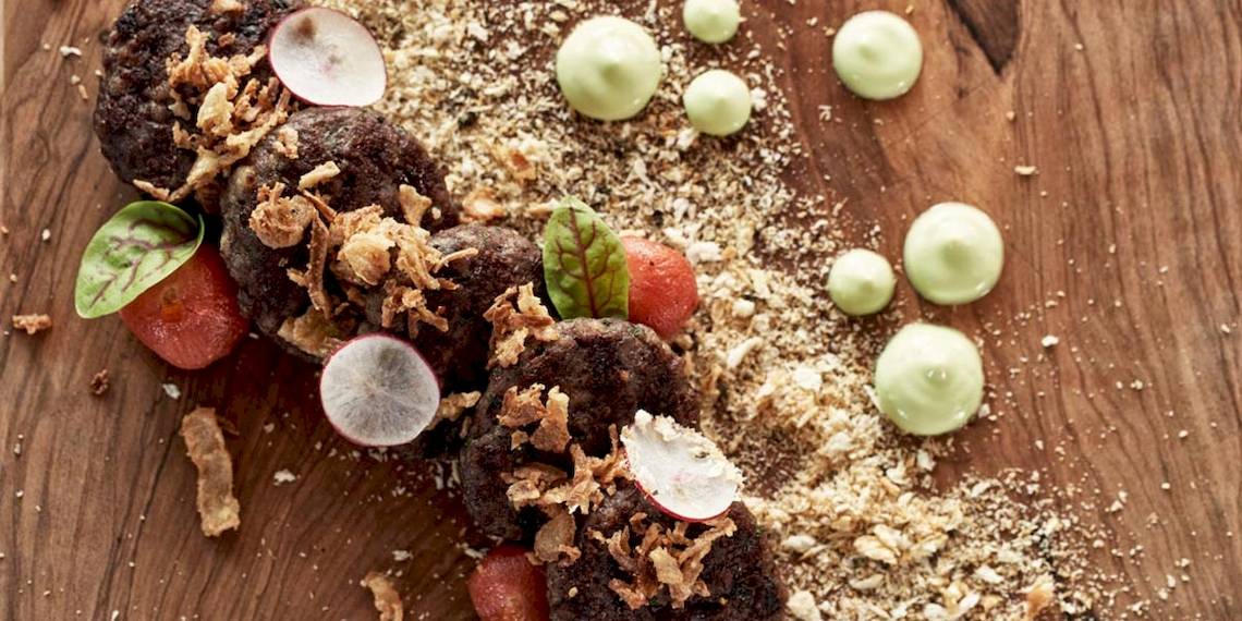 Essenplatte mit schwarzen Burgern, Radieschen, Tomaten, grünen Saucen und Krusten auf einem Holztisch.