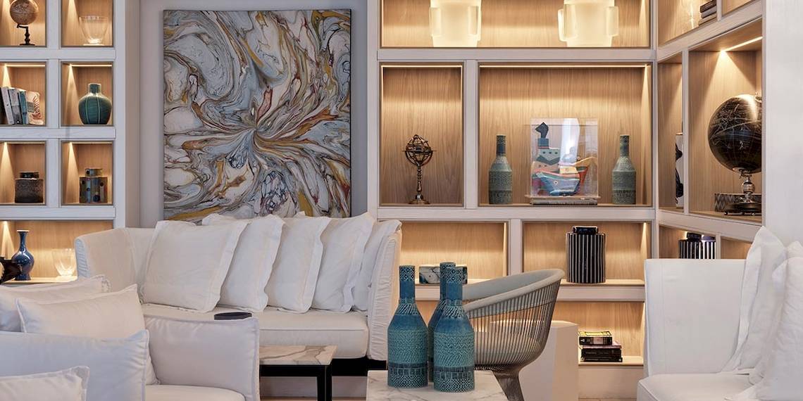 Hotellobby des Myconian Kyma mit weißen Möbeln, einer großen Wandkunst und beleuchteten Regalen.
