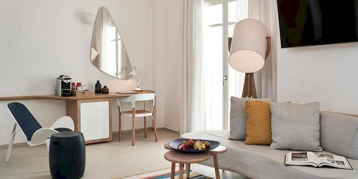 Zimmer im Myconian Kyma Hotel mit Schreibtisch, Stuhl, Lampe, Couch und offenem Buch.