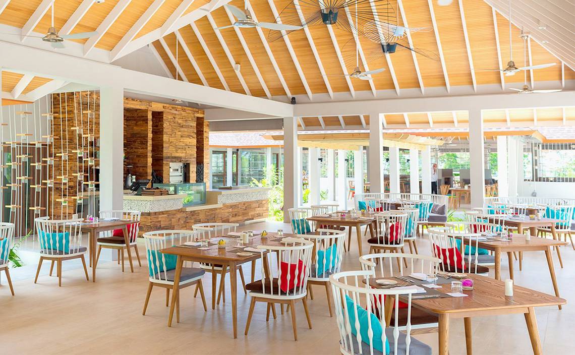 Innenbereich des Dhigali Maldives Hotels mit Holztischen, Stühlen und Deckenventilatoren.