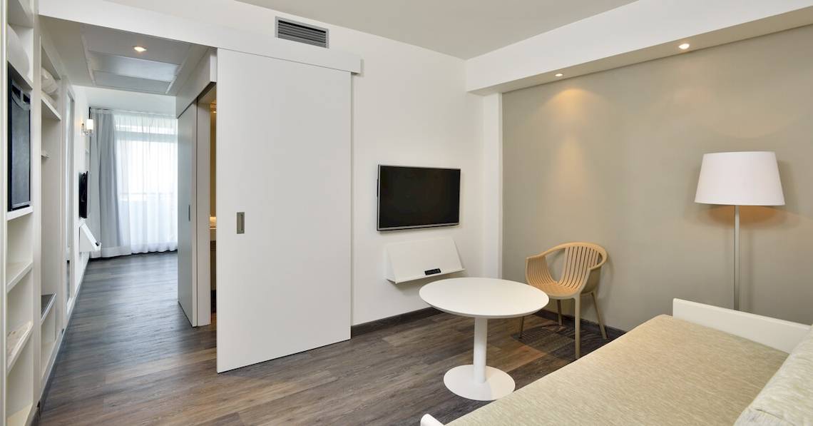 Innenansicht eines Zimmers im Sol Principe Hotel mit offenem Schiebetür, Flachbildfernseher an der Wand, weißem Tisch, Stuhl und Stehlampe.