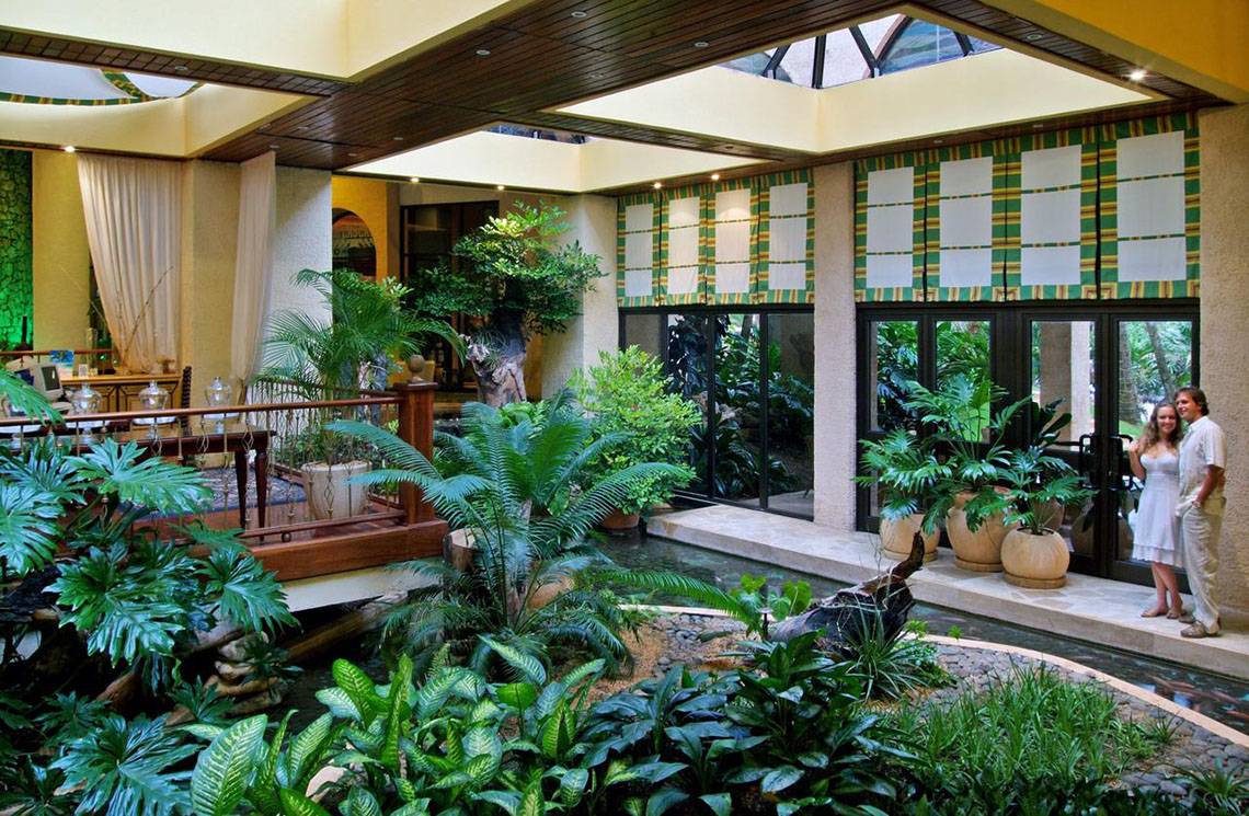 Zwei Personen stehen im Gartenbereich des Melia Las Americas Hotels, umgeben von Pflanzen und einem Wasserlauf.