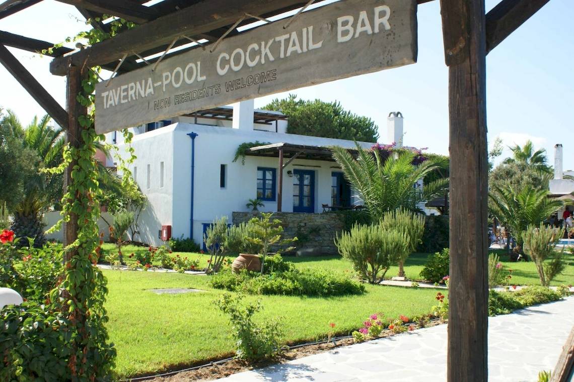 Ein Schild der Taverna-Pool-Cocktail-Bar, das vor einem weißen Hotelgebäude mit blauen Fenstern und einer Gartenlandschaft steht.