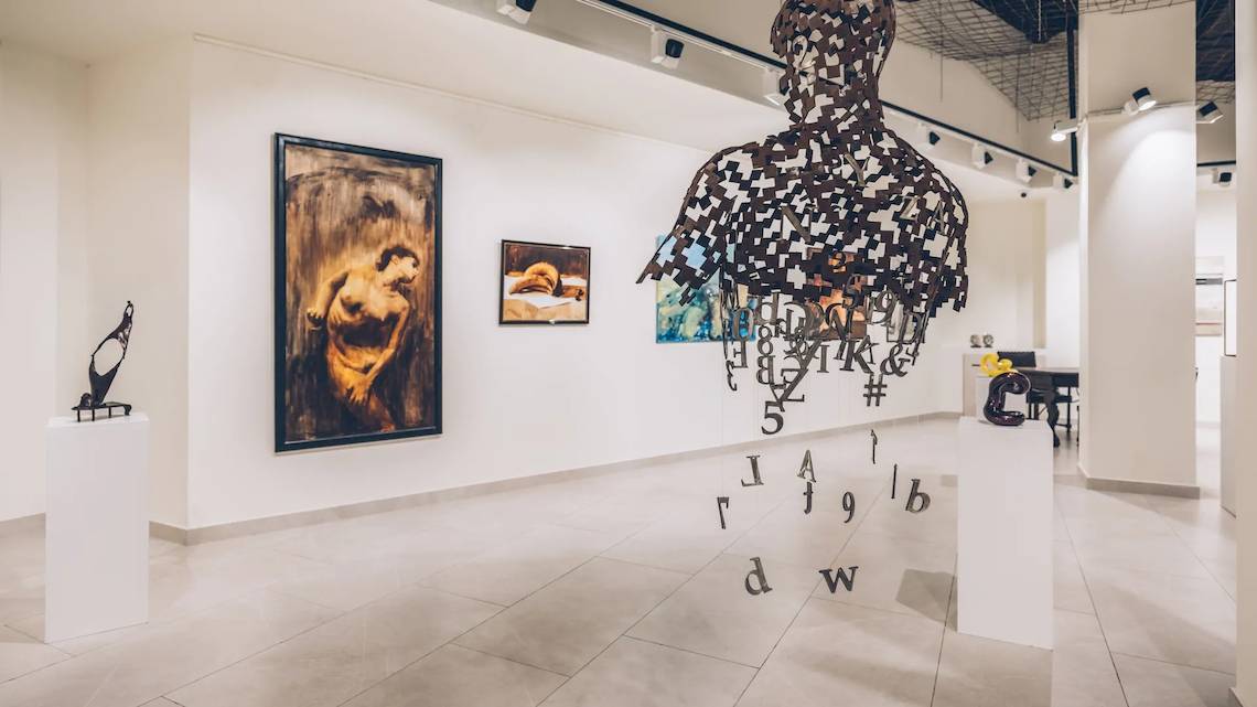 Ausstellung von Kunstwerken im Innenraum mit Gemälden an der Wand, einer Skulptur und einem großen Schriftbild.