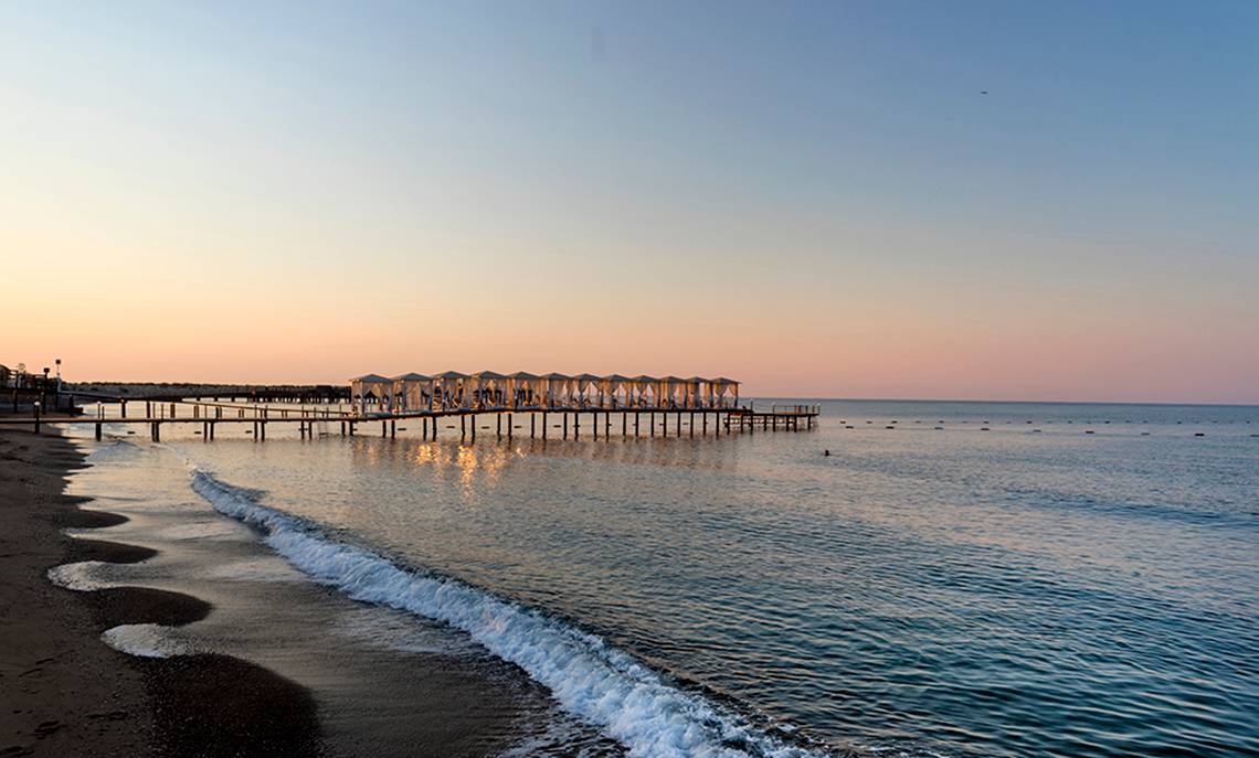 Strand mit Pier bei Sonnenuntergang, Wasser und Wellen, Gebäude im Hintergrund