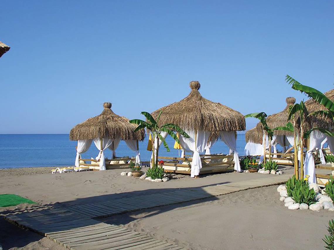 Strandszene mit mehreren Strohhütten, weißen Vorhängen und Blick auf das Meer