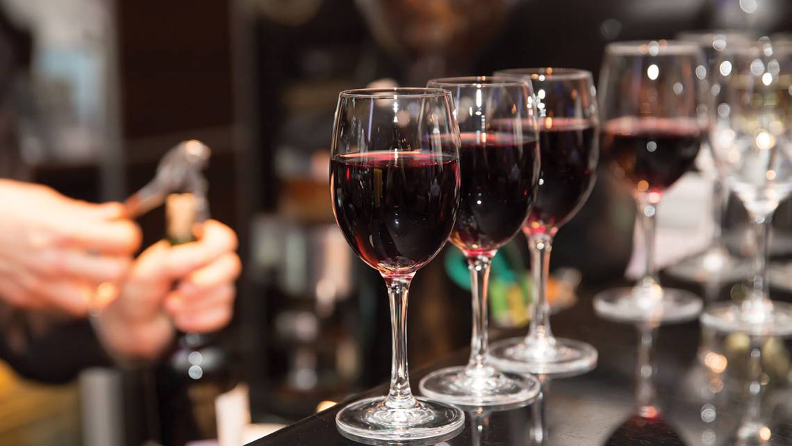 Mehrere Gläser Rotwein stehen auf einer Bar, während jemand im Hintergrund eine Flasche öffnet.