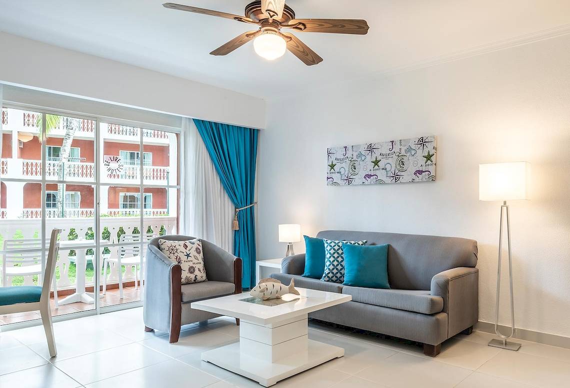 Wohnzimmer im Marien Puerto Plata Hotel mit grauem Sofa, weißem Tisch und Blick auf Balkon.