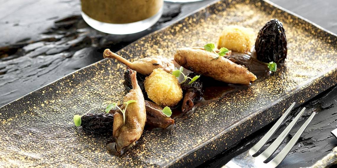 Eine goldglänzende Platte mit verschiedenen Gerichten wie gebratenen Entenbeinen, gedünsteten Früchten und Sauce, serviert im Myconian Villa Collection Hotel.