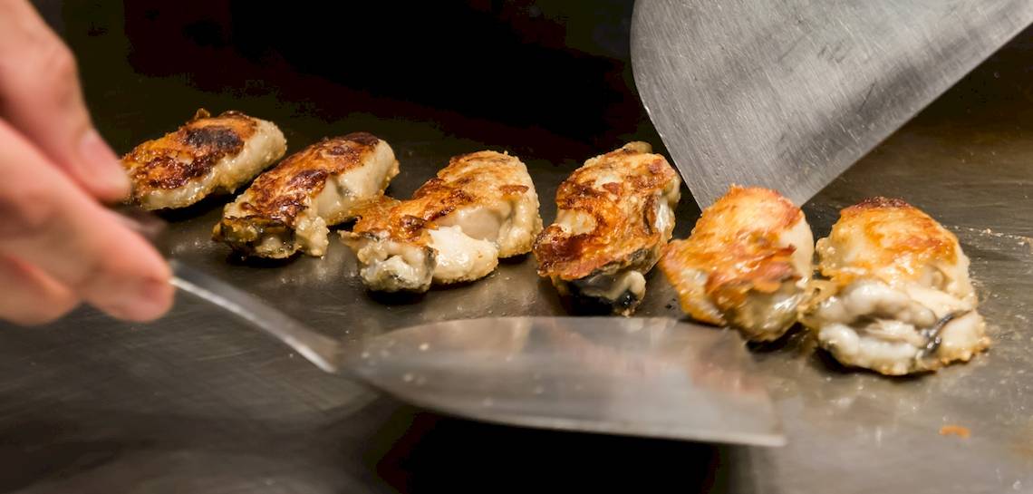 Hand schneidet gebratenes Okonomiyaki auf Grill mit mehreren Stücken
