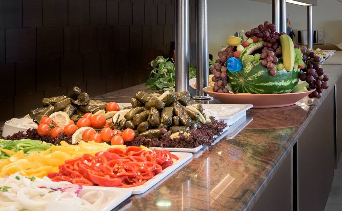 Buffet-Tisch mit verschiedenen Gemüsesorten und Obst, einschließlich Tomaten, Paprika, Salat, Auberginen und einem Wassermelonenkorb mit Früchten.