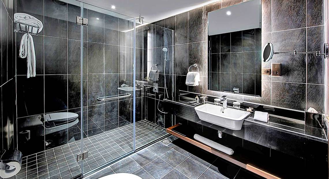 Schwarz gefliestes Badezimmer im Dukes The Palm, einem Royal Hideaway Hotel, mit einer Glasdusche, einem Waschbecken und einem Spiegel.