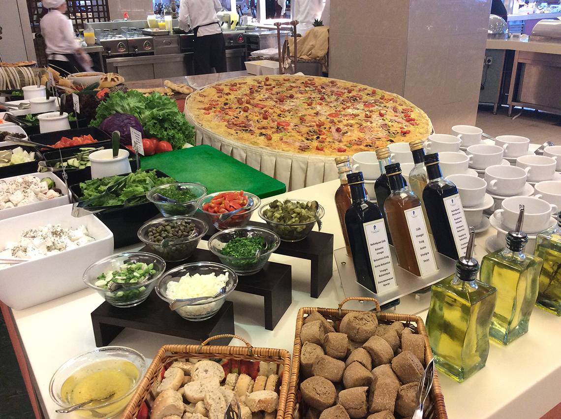 Große Pizza auf Buffettisch im Rodos Palladium Hotel mit verschiedenen Beilagen und Ölflaschen.