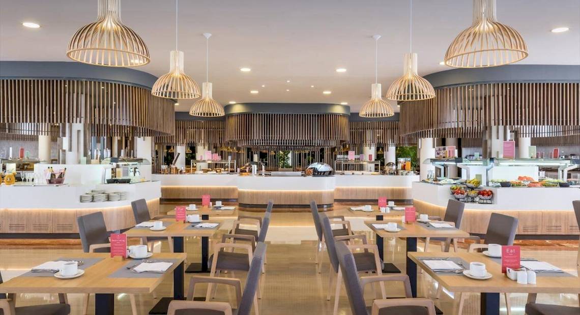 Innenansicht des Restaurants im Barcelo Corralejo Bay Hotel mit geometrischen Pendelleuchten, hölzernen Elementen und gedeckten Tischen.