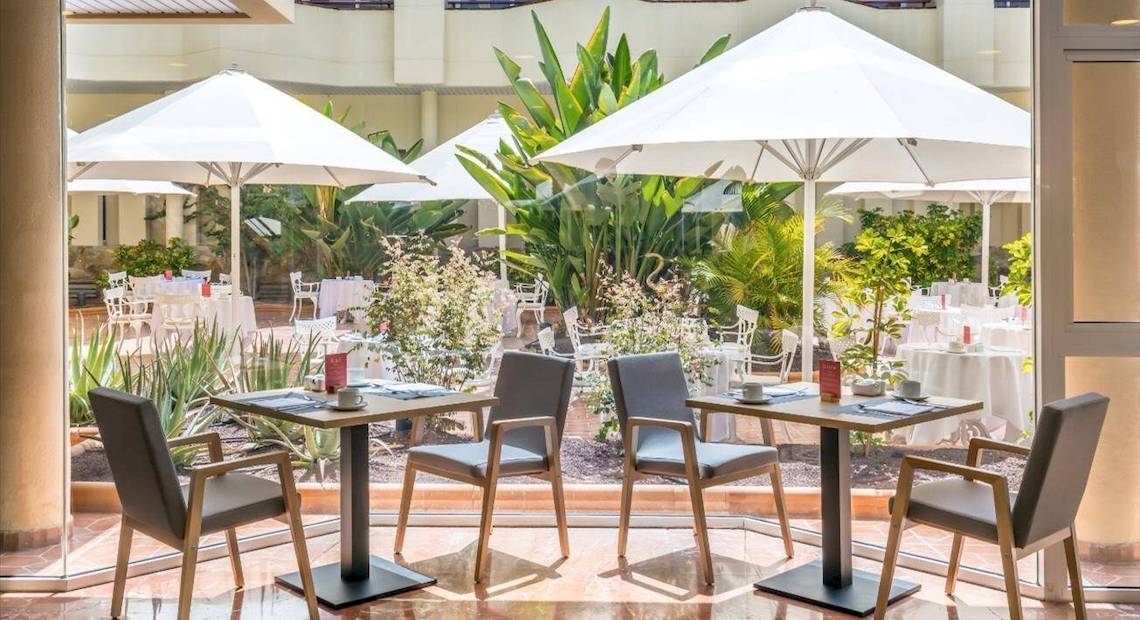 Gartenbereich des Barcelo Corralejo Bay Hotels mit Tischen, Stühlen und Schirmen.