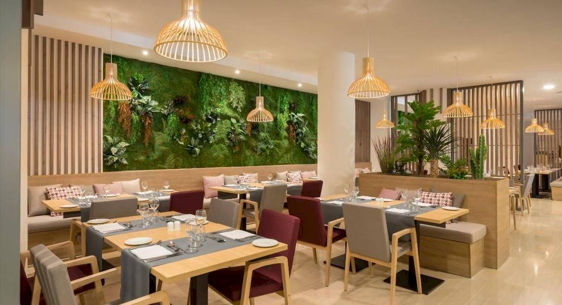 Innenbereich des Restaurants des Barcelo Corralejo Bay Hotels mit grüner Wand, Holztischen und Stühlen.