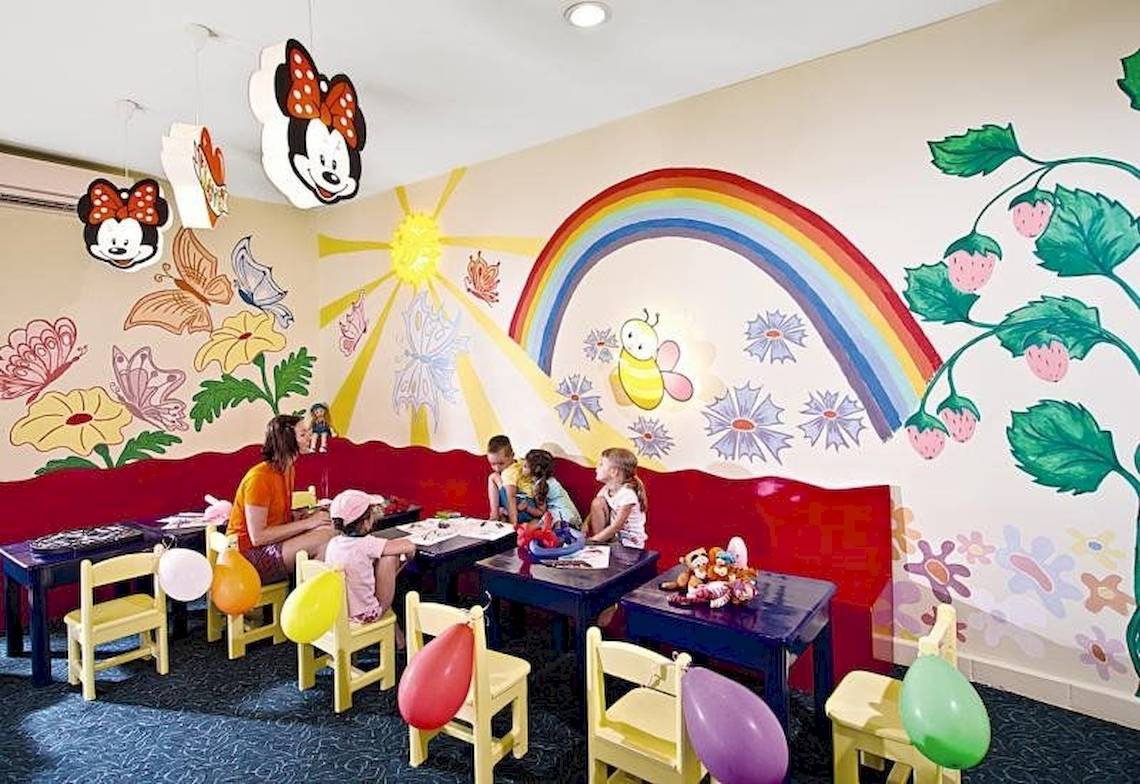 Spielzimmer im Serenity Alma Heights Hotel mit bunten Wandmalereien, bunten Stühlen und Tischen, Luftballons und einer Frau, die Kindern hilft.