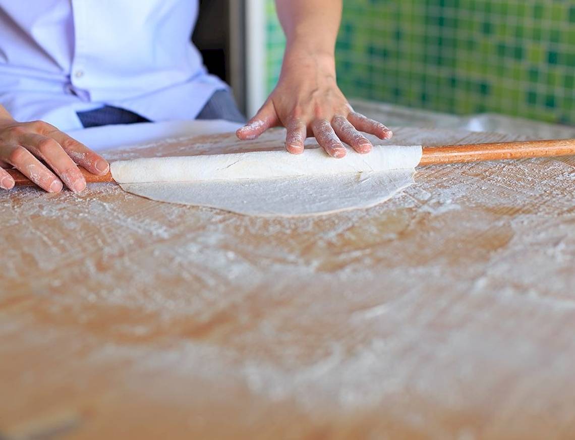 Person rollt Teig mit einer Holzrolle auf einem bemehlten Tisch.