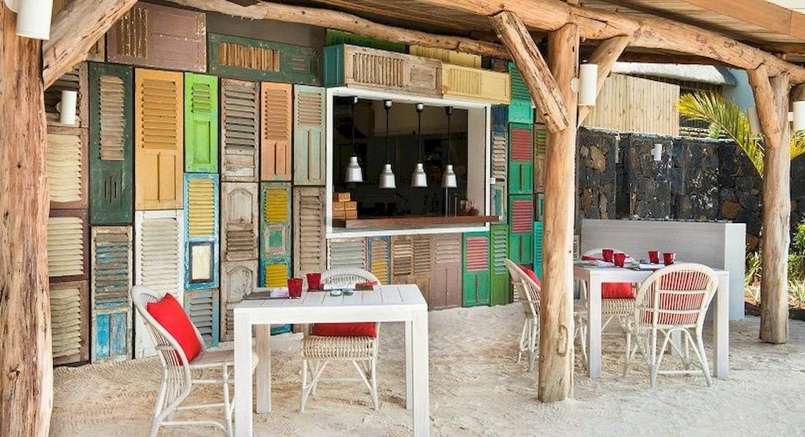 Außenbereich des Zilwa Attitude Hotels mit bunten Fensterläden und Tischen und Stühlen auf weißem Sand.
