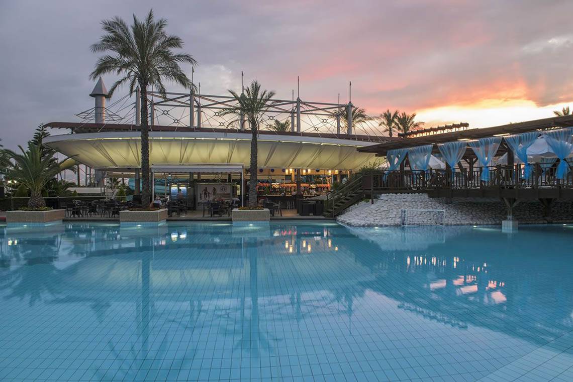 Außenpool des Delphin Diva Premiere Hotels bei Sonnenuntergang mit blauem Himmel und Palmen.