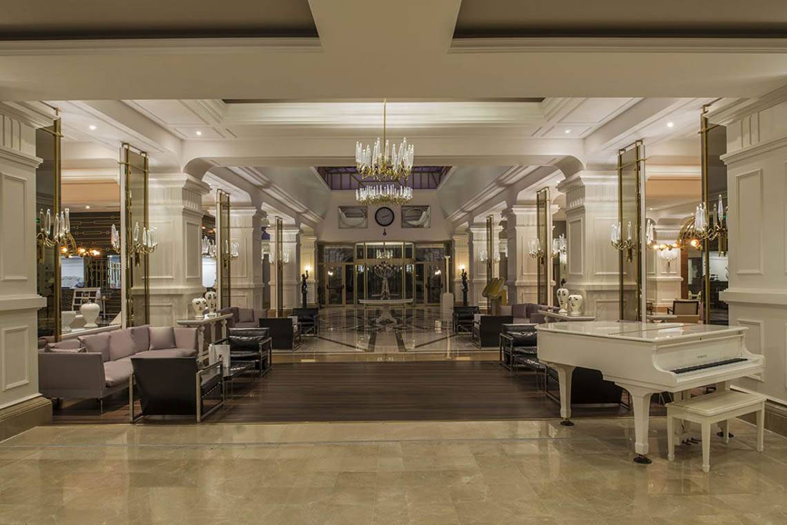 Das elegante Foyer des Delphin Diva Premiere Hotels mit einem weißen Klavier, Sofas und Kronleuchtern.