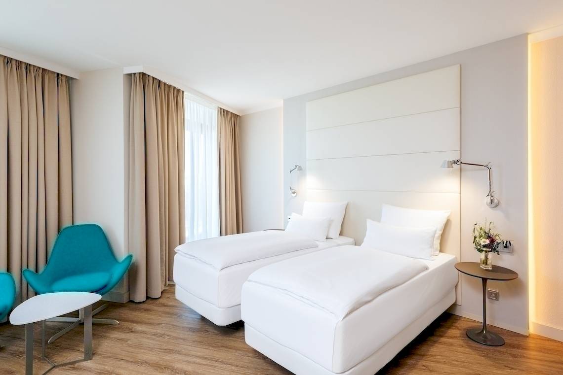 Zimmer im NH Berlin Alexanderplatz Hotel mit zwei weißen Betten und modernen Möbeln.