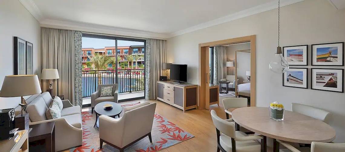 Innenansicht der Suite im Hilton Cabo Verde Sal Resort mit Wohnzimmer, Esstisch, Fernseher und Balkon.