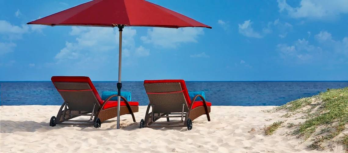 Zwei rote Sonnenliegen mit Kissen und einem roten Sonnenschirm auf dem Strand am Hilton Cabo Verde Sal Resort.