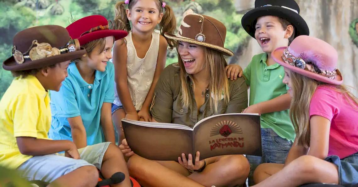Eine Frau mit Hut liest ein Buch vor, während mehrere Kinder, ebenfalls mit Hüten, ihr zuhören.