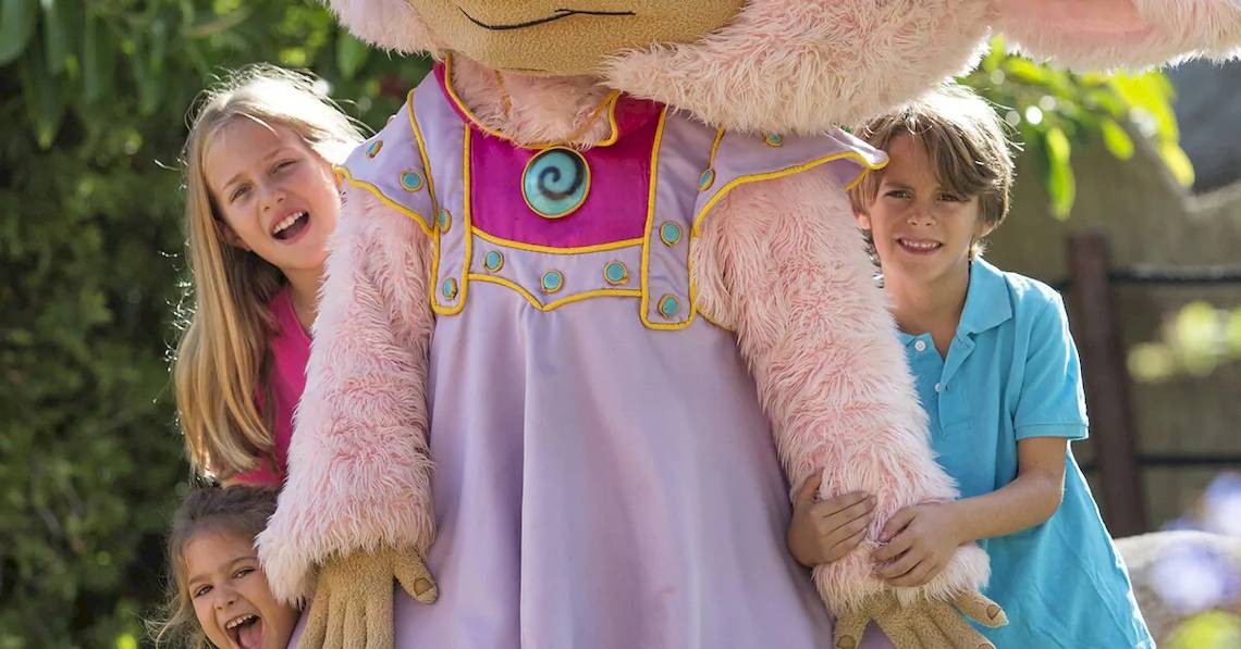 Drei Kinder, zwei Mädchen und ein Junge, posieren mit einem pinken Mädchen Maskottchen in einem Garten.
