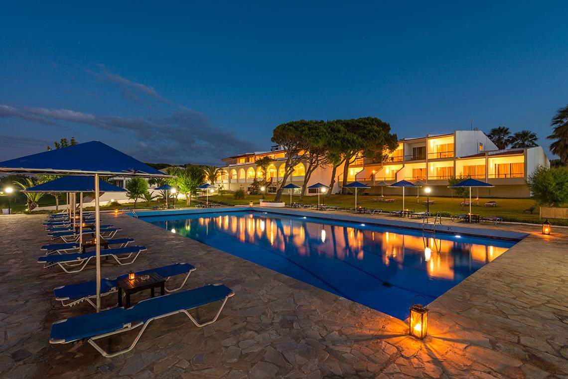 Abendlicher Blick auf den Pool im Pavlina Beach Hotel mit blauen Sonnenschirmen und Lounge-Liegestühlen.