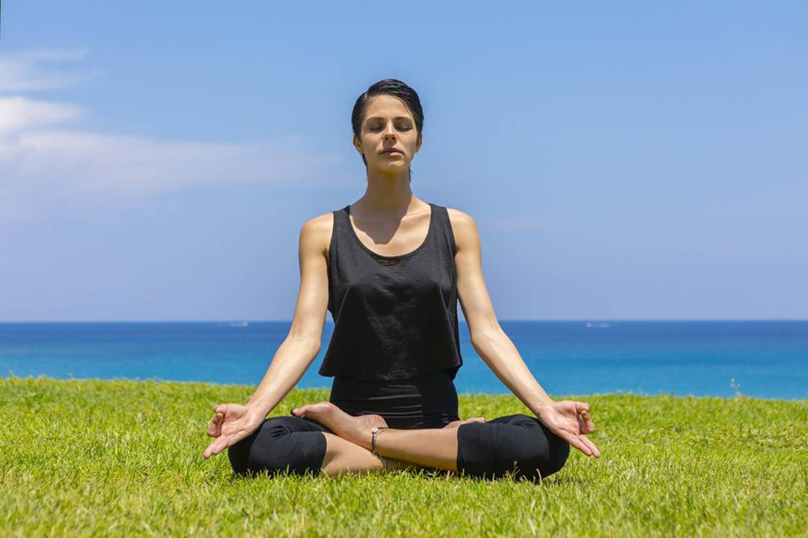 Frau im Yogasitz auf dem Rasen vor dem Meer am Strand des Cavo Maris Beach Hotels