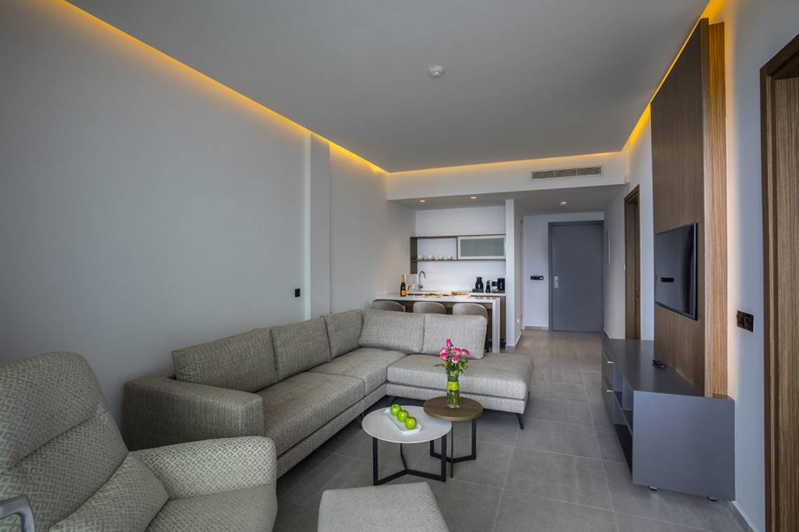 Wohnzimmer im Cavo Maris Beach Hotel mit L-förmiger Couch, Tisch, Fernseher und Küche im Hintergrund.