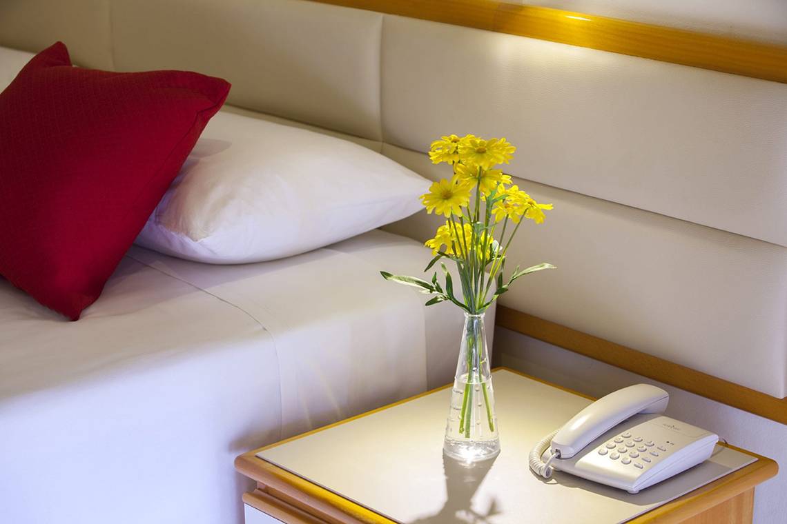 Ein Hotelzimmer mit einem Bett, zwei Kissen, einer Vase mit gelben Blumen und einem Telefon auf dem Nachttisch.