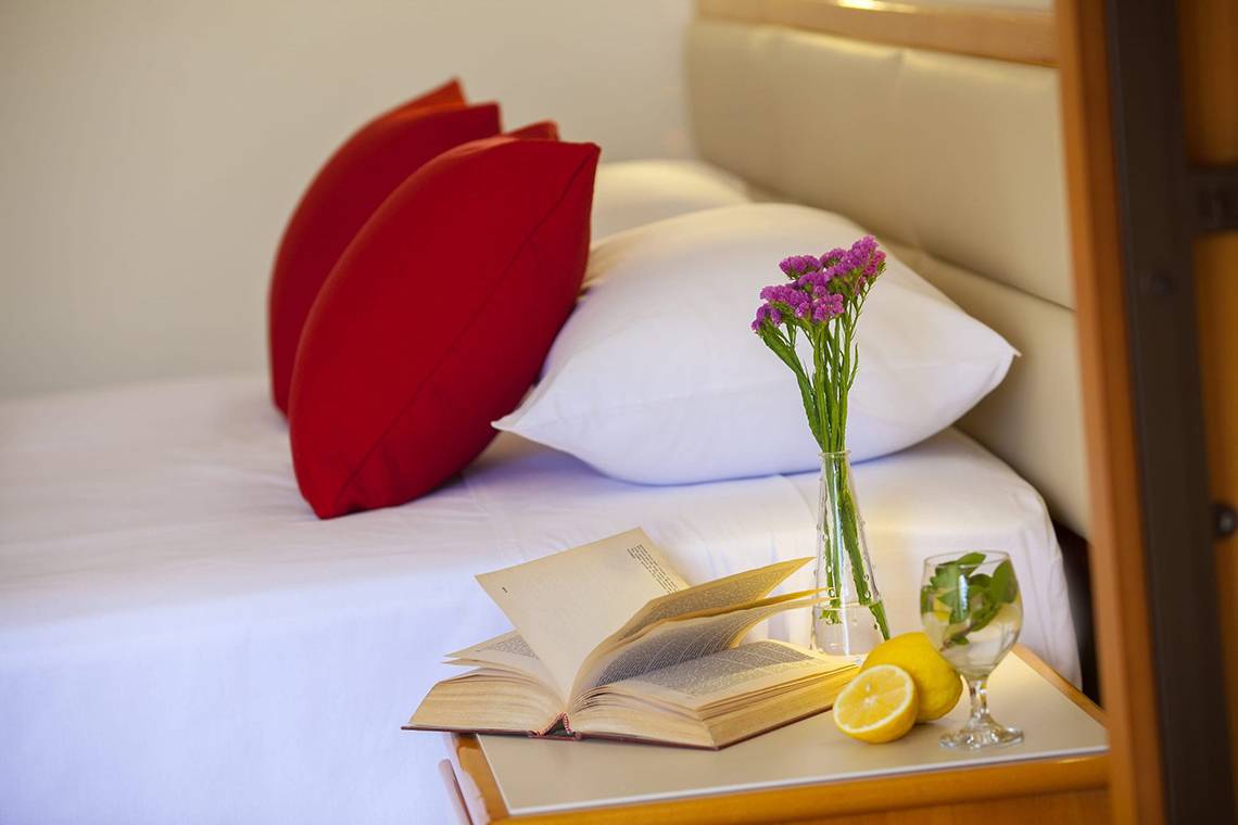 Ein Bett im Queens Bay Hotel mit roten Kissen, einer offenen Buche, einer Vase mit Blumen, einem Glas mit Limonade und Zitronen.
