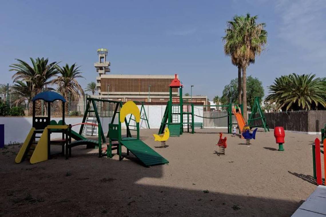 Ein Kinderspielplatz im Freien mit verschiedenen Spielgeräten, Palmen und einem Gebäude im Hintergrund.