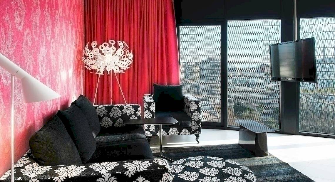 Wohnzimmer im Barcelo Raval Hotel mit schwarzem und weißem Sofa, roter Vorhang, Tisch, Fernseher und Lampen.