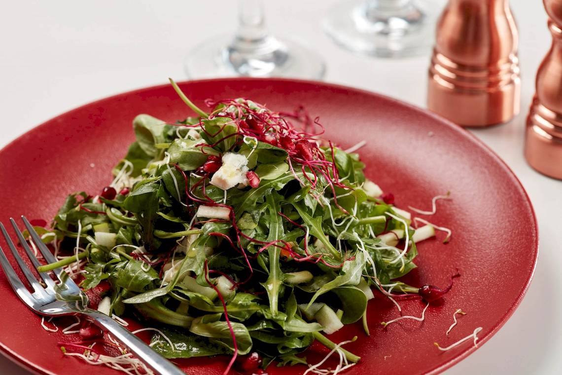 Ein roter Teller mit einem gemischten Salat, der mit Feta-Käse und Granatapfel-Samen garniert ist, und eine Gabel.
