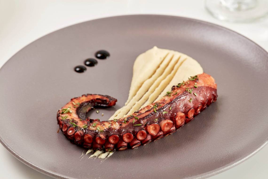Ein elegantes Gericht mit einem gegrillten Oktopus auf einem Teller mit Beilagen und Sauce, im Hotel Absolute Mykonos Suites & More serviert.