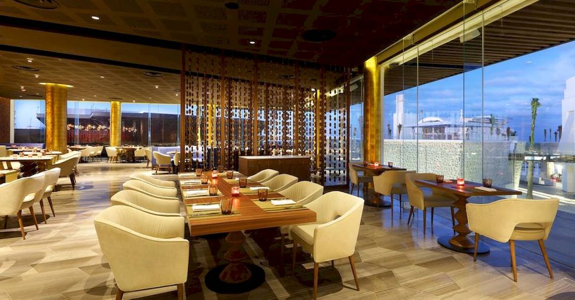 Innenansicht des Restaurants im Grand Palladium Costa Mujeres Resort mit runden Tischen, weißen Stühlen und großen Fenstern.