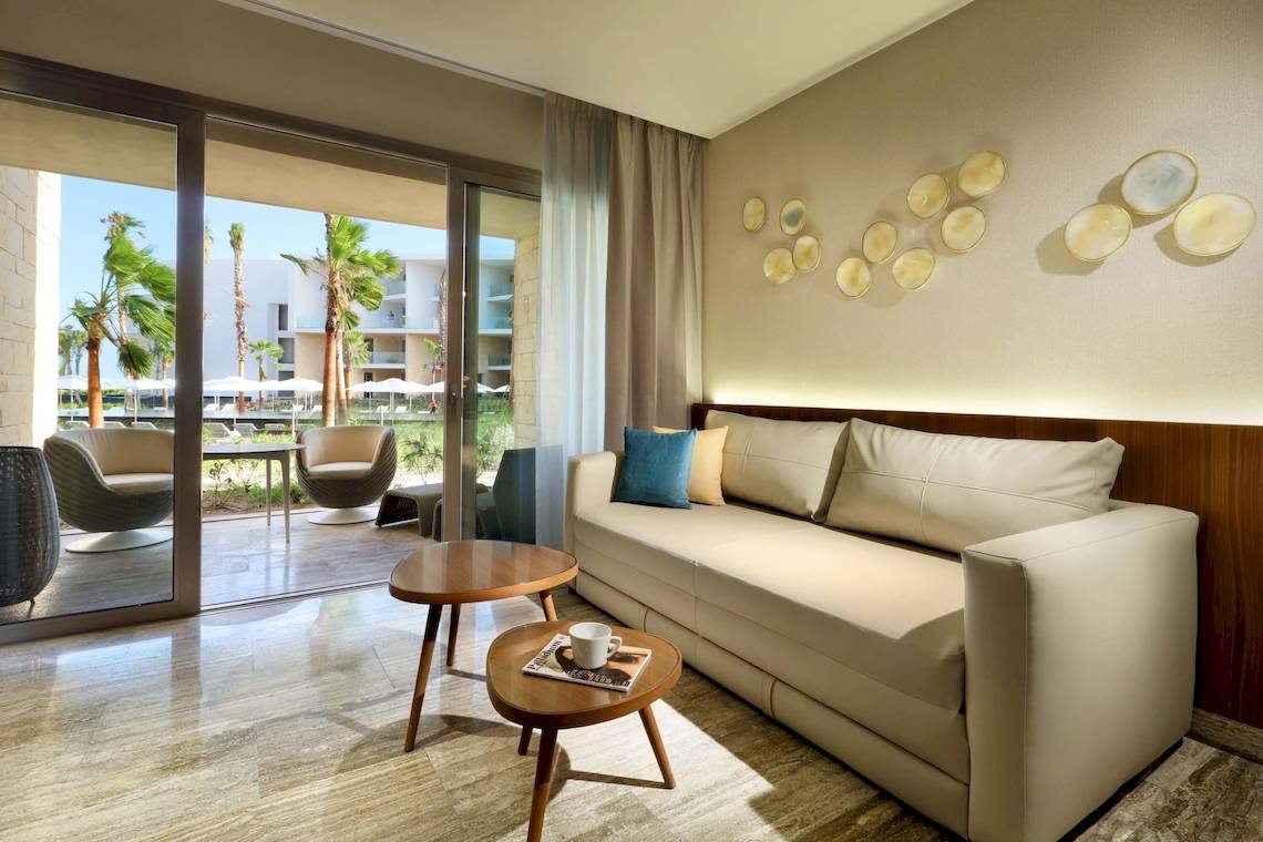 Wohnbereich im Grand Palladium Costa Mujeres Resort & Spa mit einer Couch, zwei Tischen und einer Aussicht auf Palmen und Gebäude.