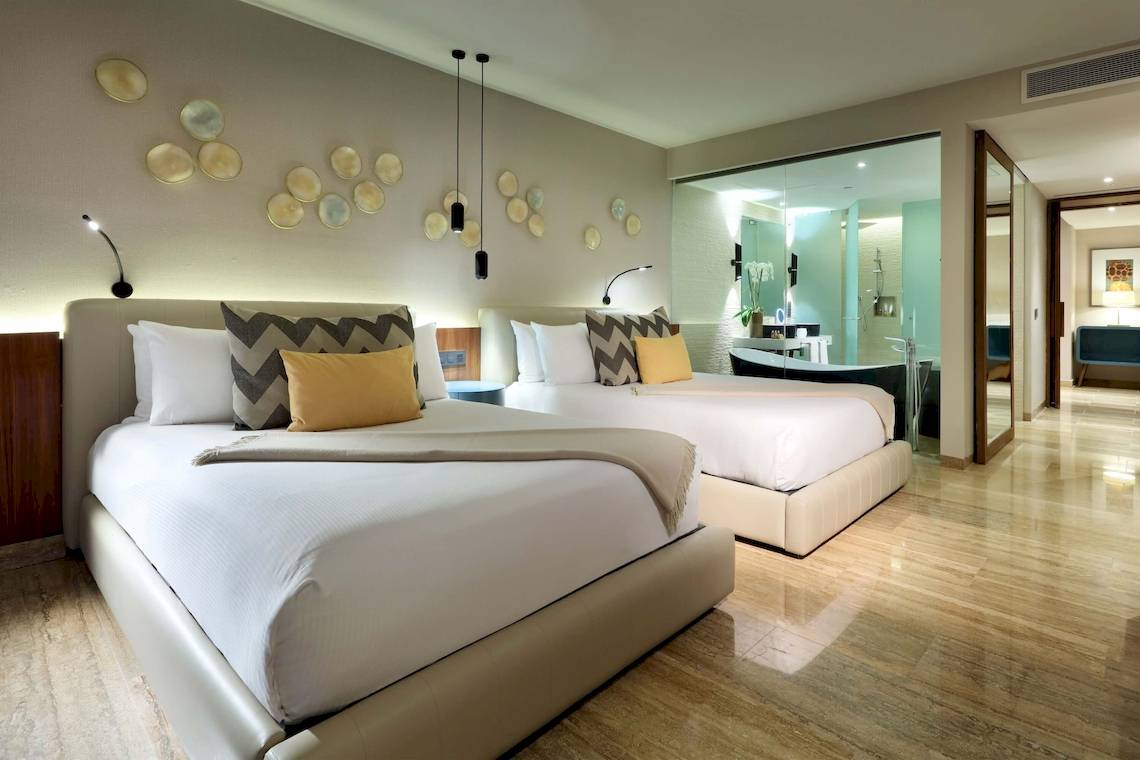 Zwei Betten im Grand Palladium Costa Mujeres Resort & Spa mit weißen Laken, Kissen und einem modernen Badezimmer.