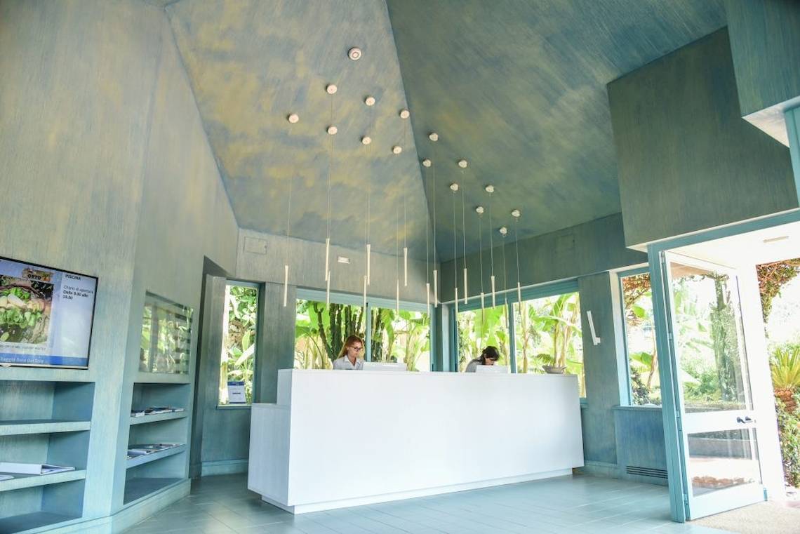Empfangsbereich des Baia del Sole Hotels mit weißen Schreibtischen, blauen Wänden und Pflanzen im Hintergrund.