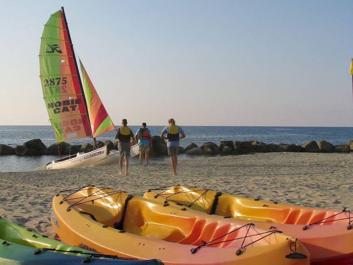 Segelboot am Strand von Baia del Sole mit drei Personen in Schwimmwesten und Kajaks auf dem Sand.