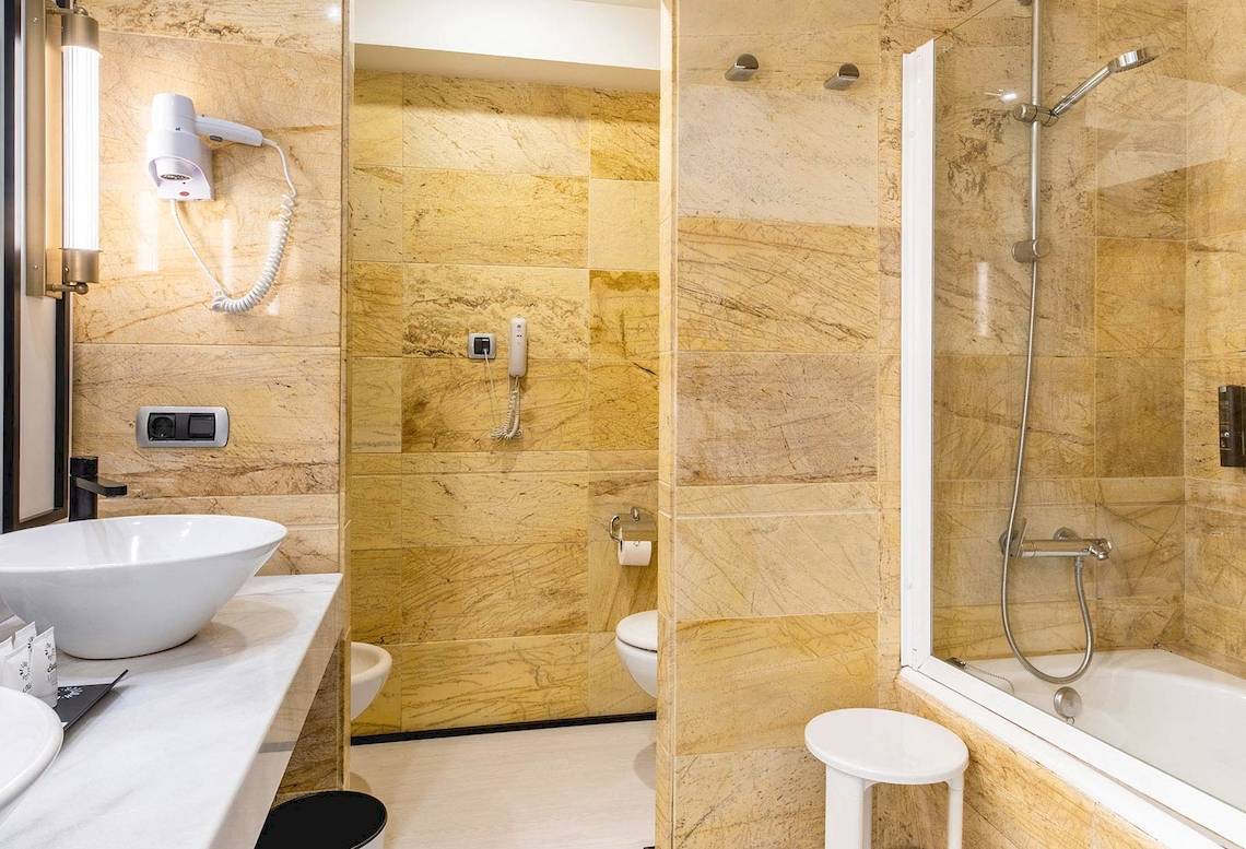 Badezimmer im Sarena de Muro Resort Mallorca mit Marmorwänden, Badewanne, Toilette und Föhn.